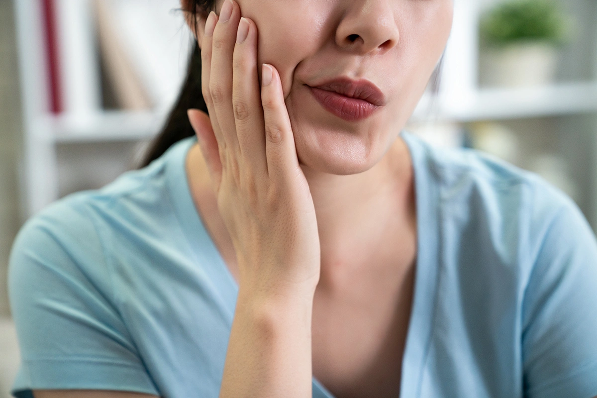 Asian woman holding her cheek with a pained expression, illustrating toothache or dental discomfort caused by sensitivity, decay or gum issues.